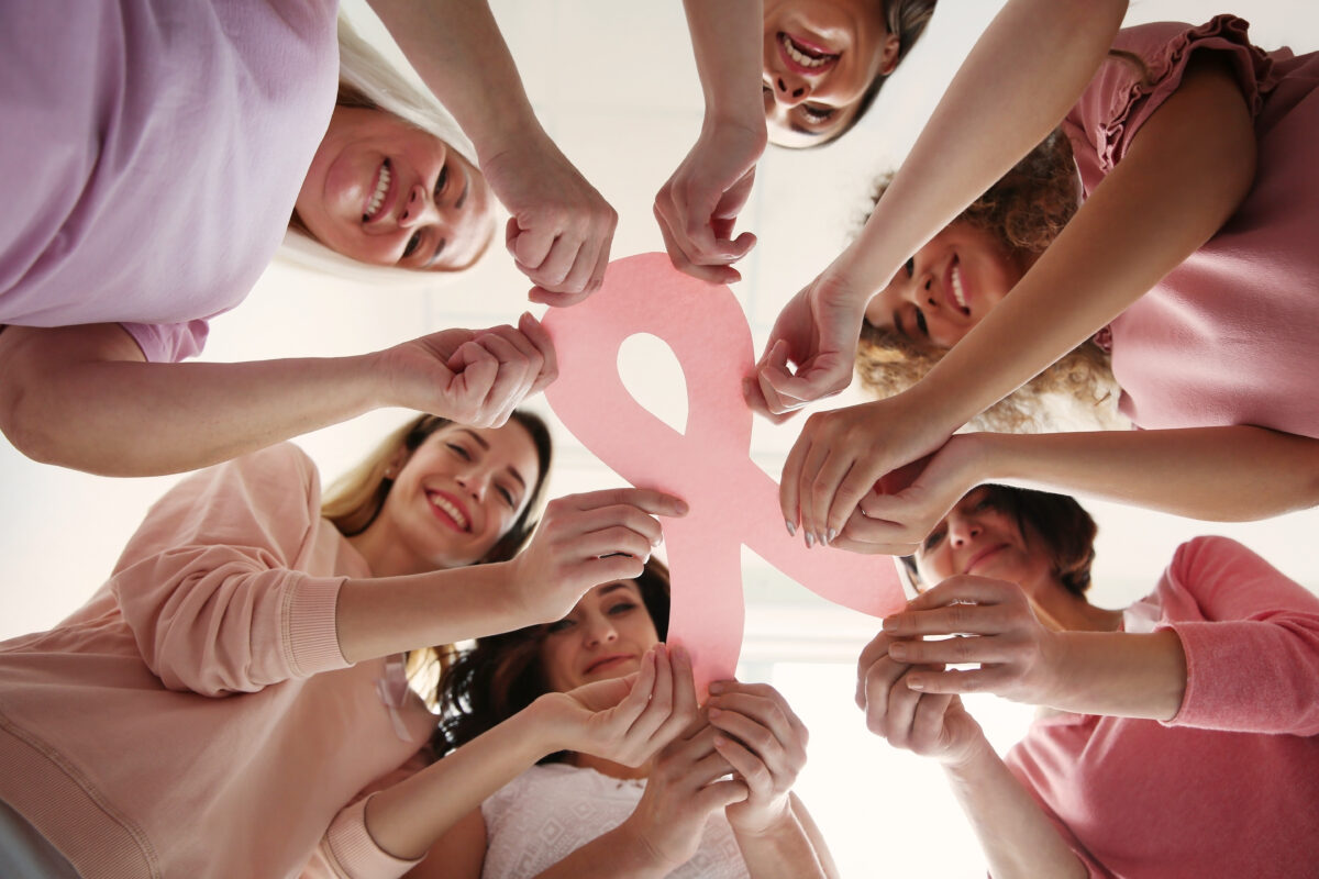 Sechs Frauen blicken nach unten und stehen im Kreis, sie halten gemeinsam eine rosa Schleife aus Papier