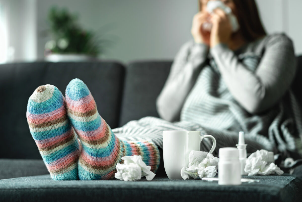 Frau sitzt krank mit Decke auf dem Sofa, trägt bunte Wollsocken, trinkt Tee, umgeben von Taschentüchern und Medikamenten.
